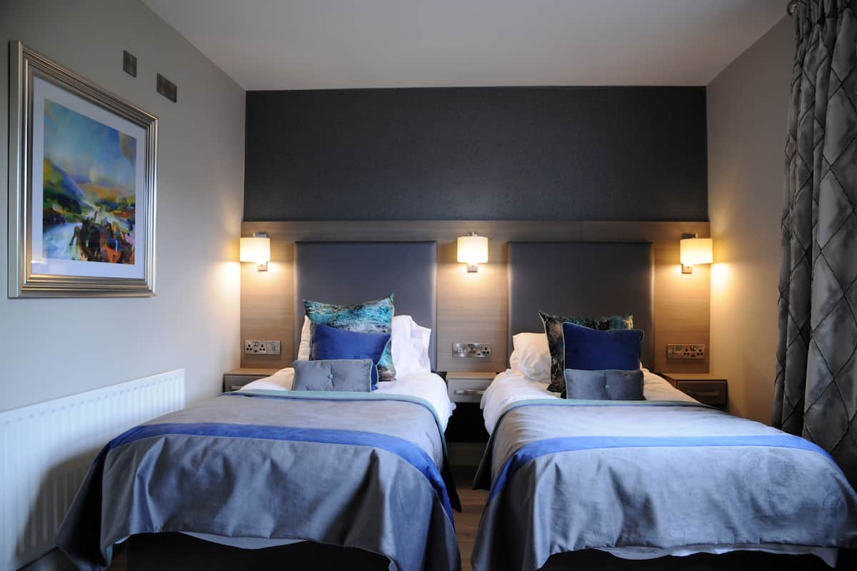 Quarto de hotel com duas camas, cada uma com colcha azul e branca, um quadro na parede e luminárias fixadas na parede.
