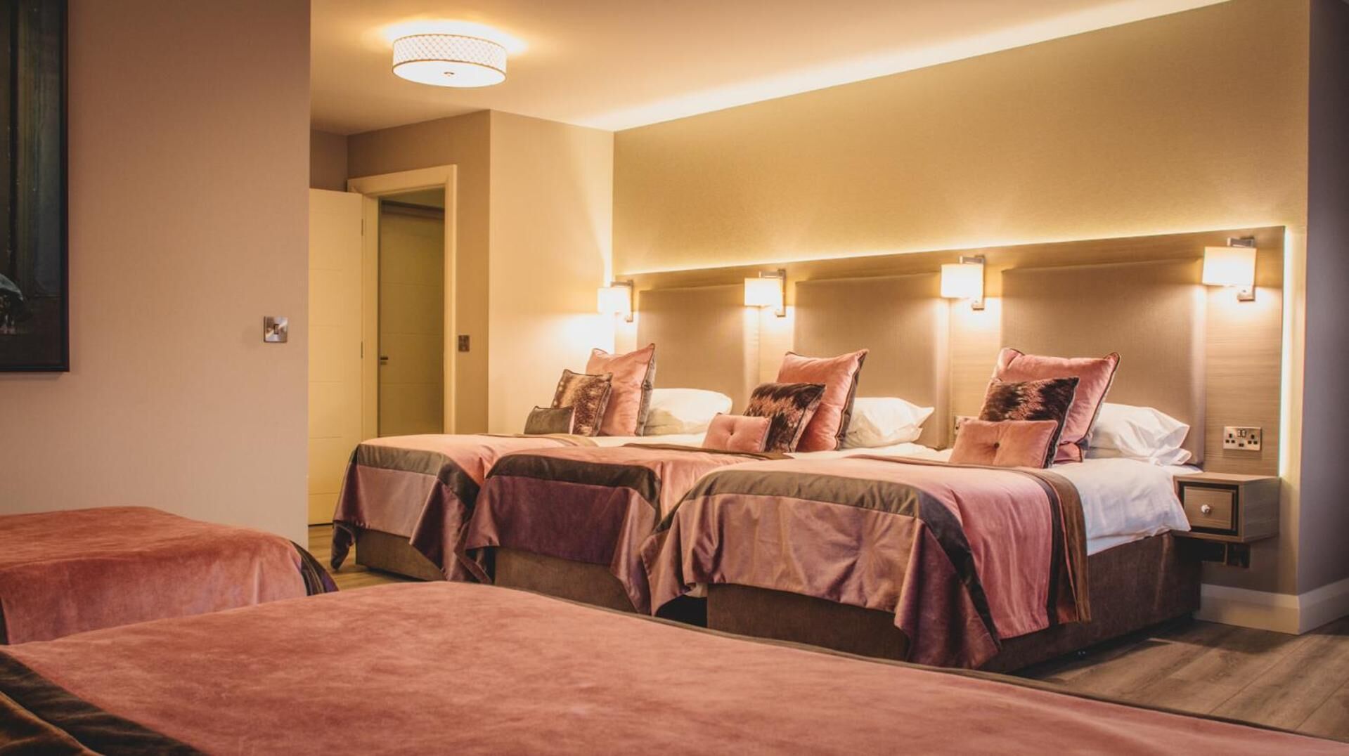 A hotel room with two beds, each with a headboard and bedside lamps. There is a door leading to a hallway.