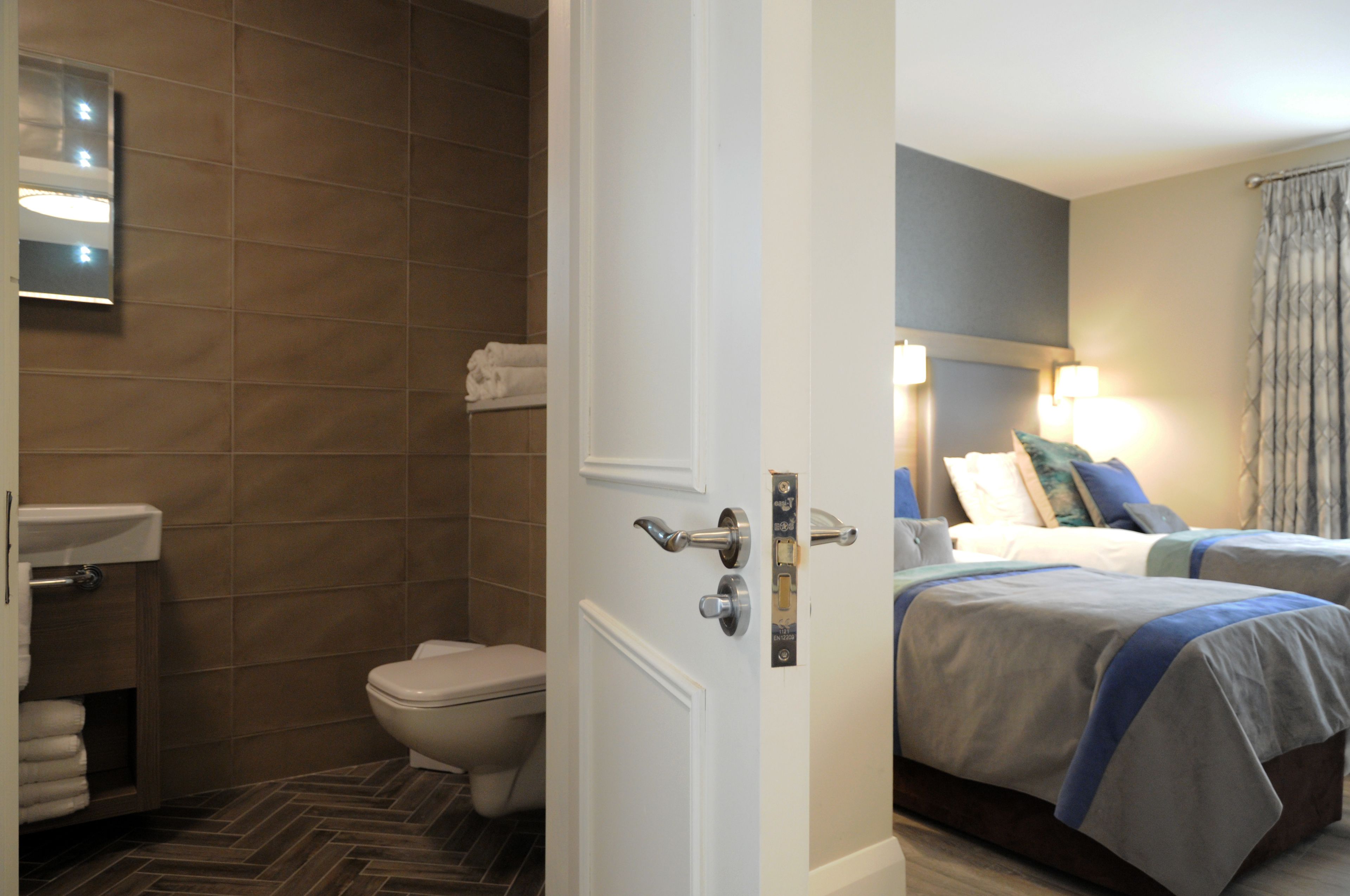 A hotel room with a bathroom, a bed with a blue blanket, and a nightstand with a lamp.