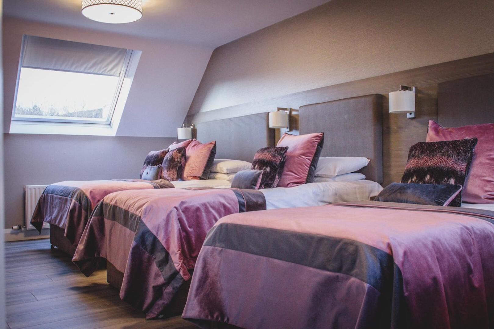 Habitación de hotel con tres camas, cada una con cabecera y lámparas de noche. Una ventana ofrece luz natural y vistas al exterior.