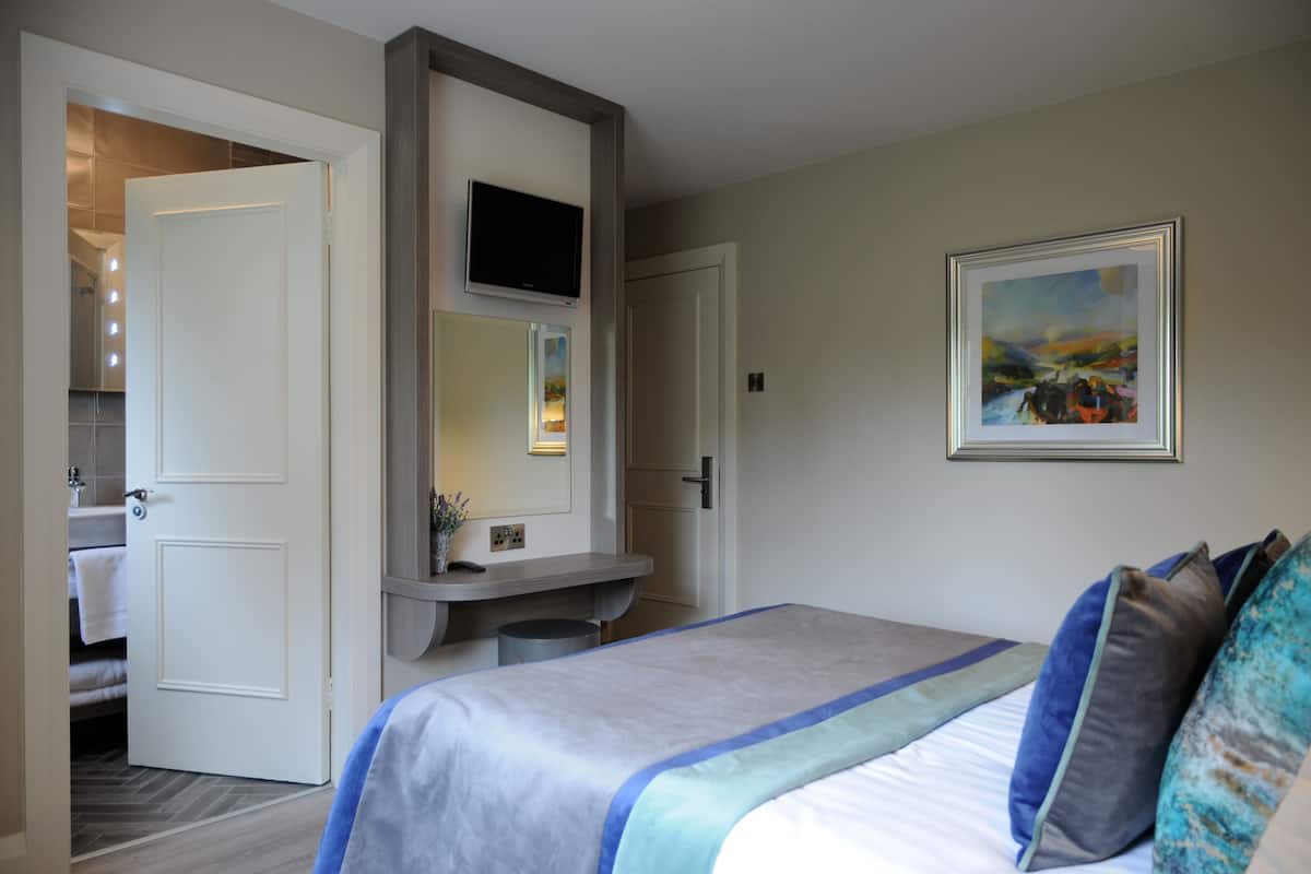 Quarto de hotel com cama, televisão, pintura e banheiro visível através de uma porta aberta.