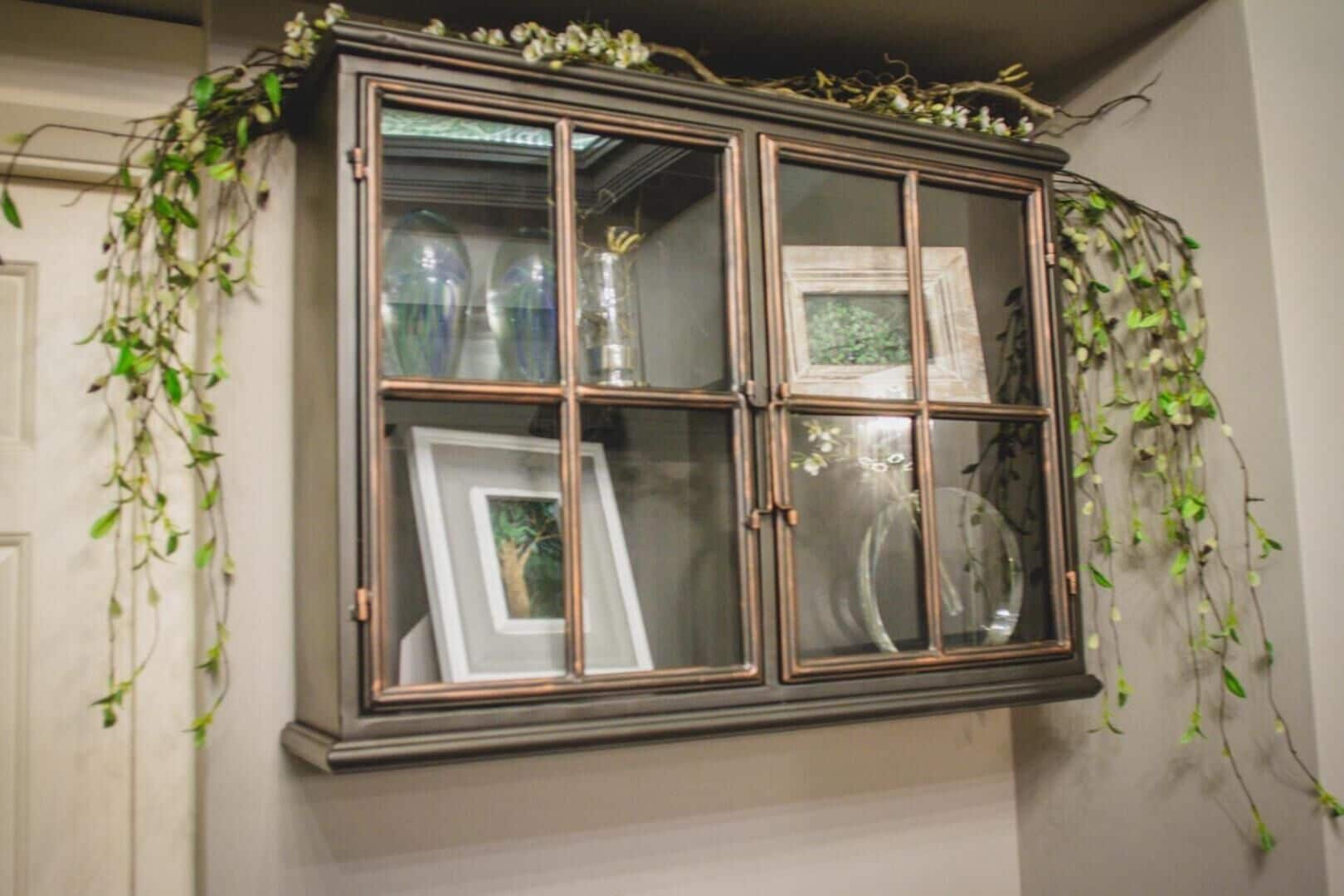 Un aparador de madera oscura con puertas de cristal, decorado con enredaderas colgantes, que contiene fotografías enmarcadas y cristalería.