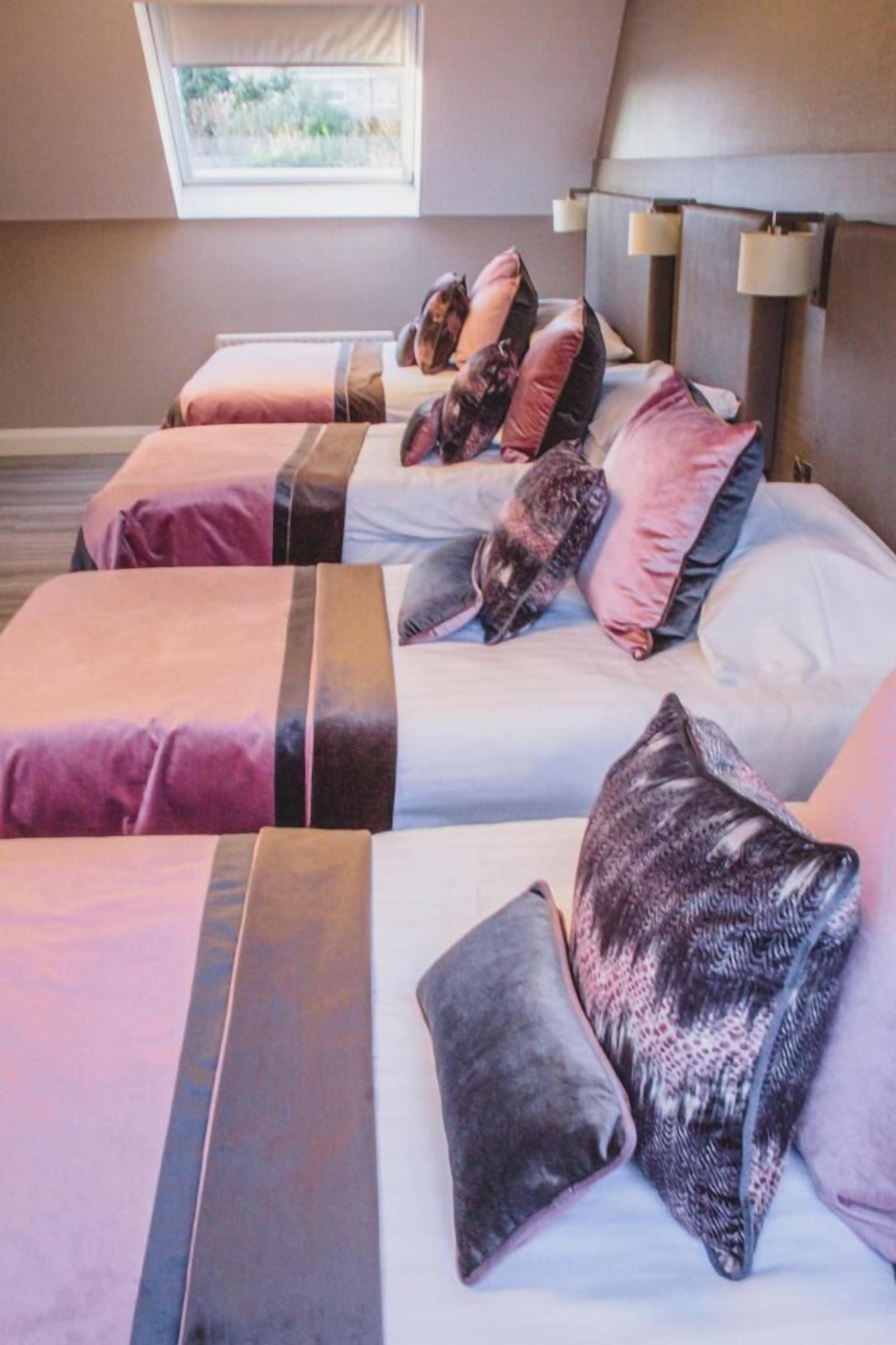 A hotel room with three beds, each with a different color bedspread and multiple pillows. There is a window with a view of greenery outside.