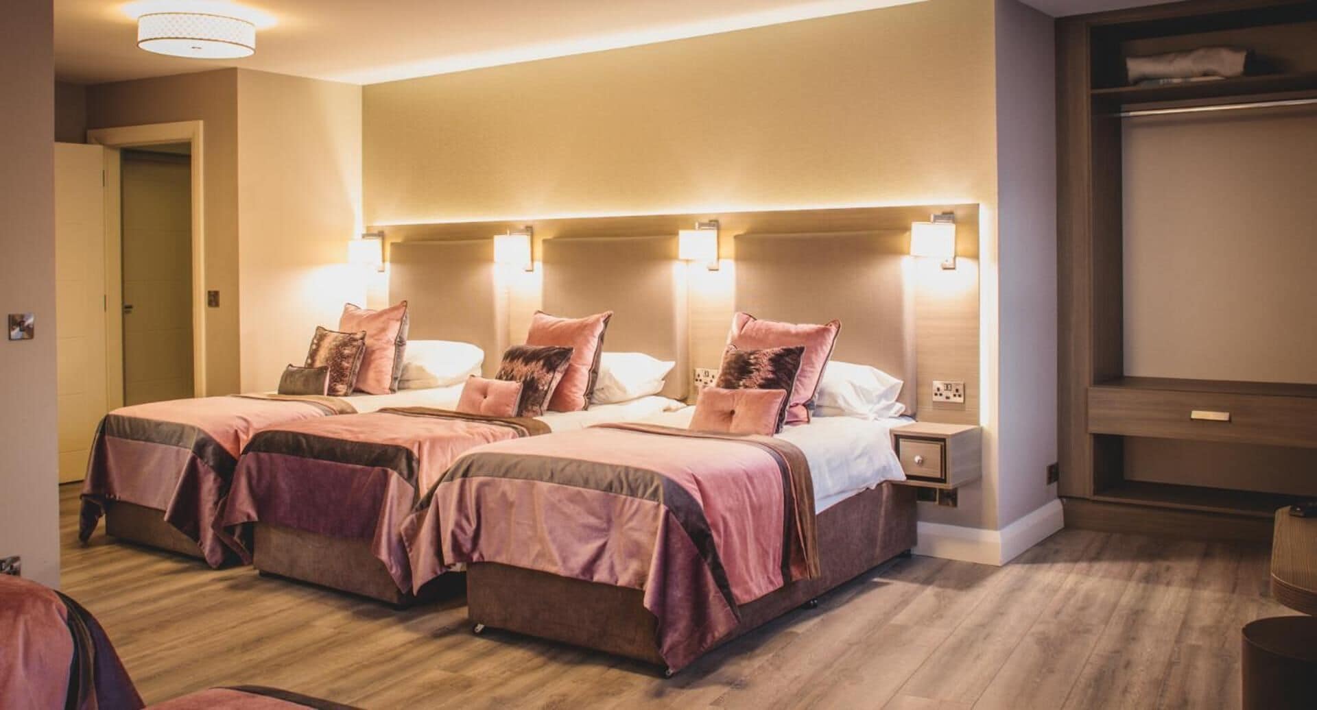 A hotel room with two beds, each with a headboard and bedside lamps. There is a wooden floor and a wardrobe in the corner.