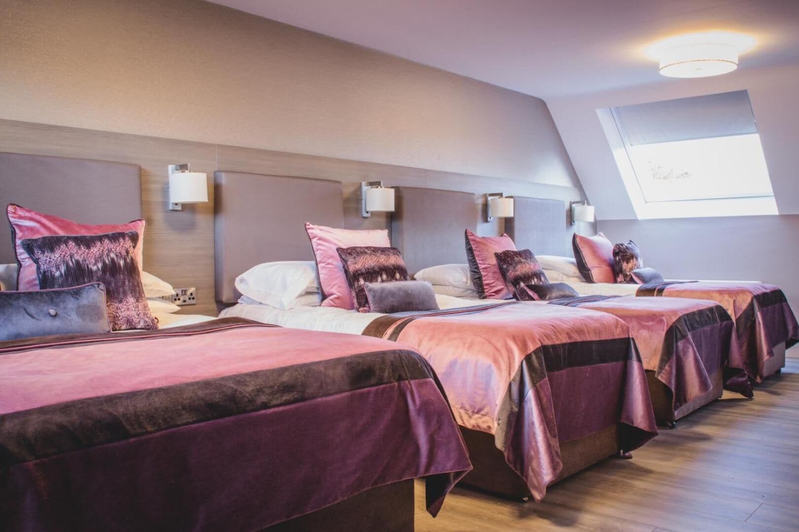Habitación de hotel con cuatro camas dispuestas en fila, cada una con cabecera y lámparas de noche.