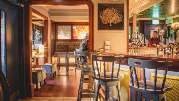 Een bar met houten barkrukken, een toog met verschillende biertaps, en een zithoek met tafels en stoelen.
