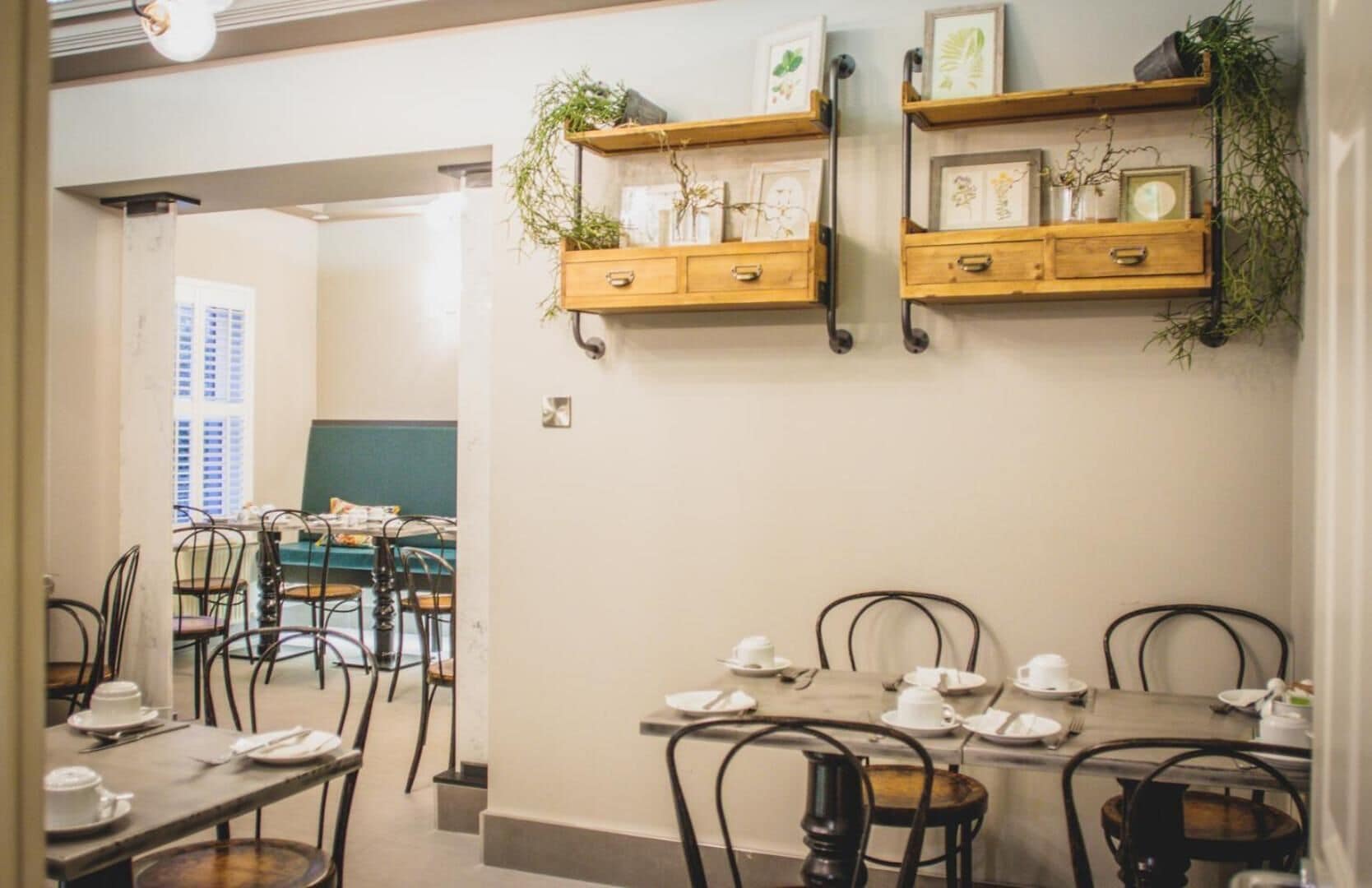 A dining area with wooden tables and chairs, some with white tableware. There are two wooden shelves with plants and framed pictures.