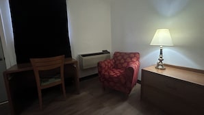 A small room with a wooden desk, a red patterned chair, a lamp, and an air conditioning unit. - Candlelight Inn by Capital O Scottsbluff (Scottsbluff)