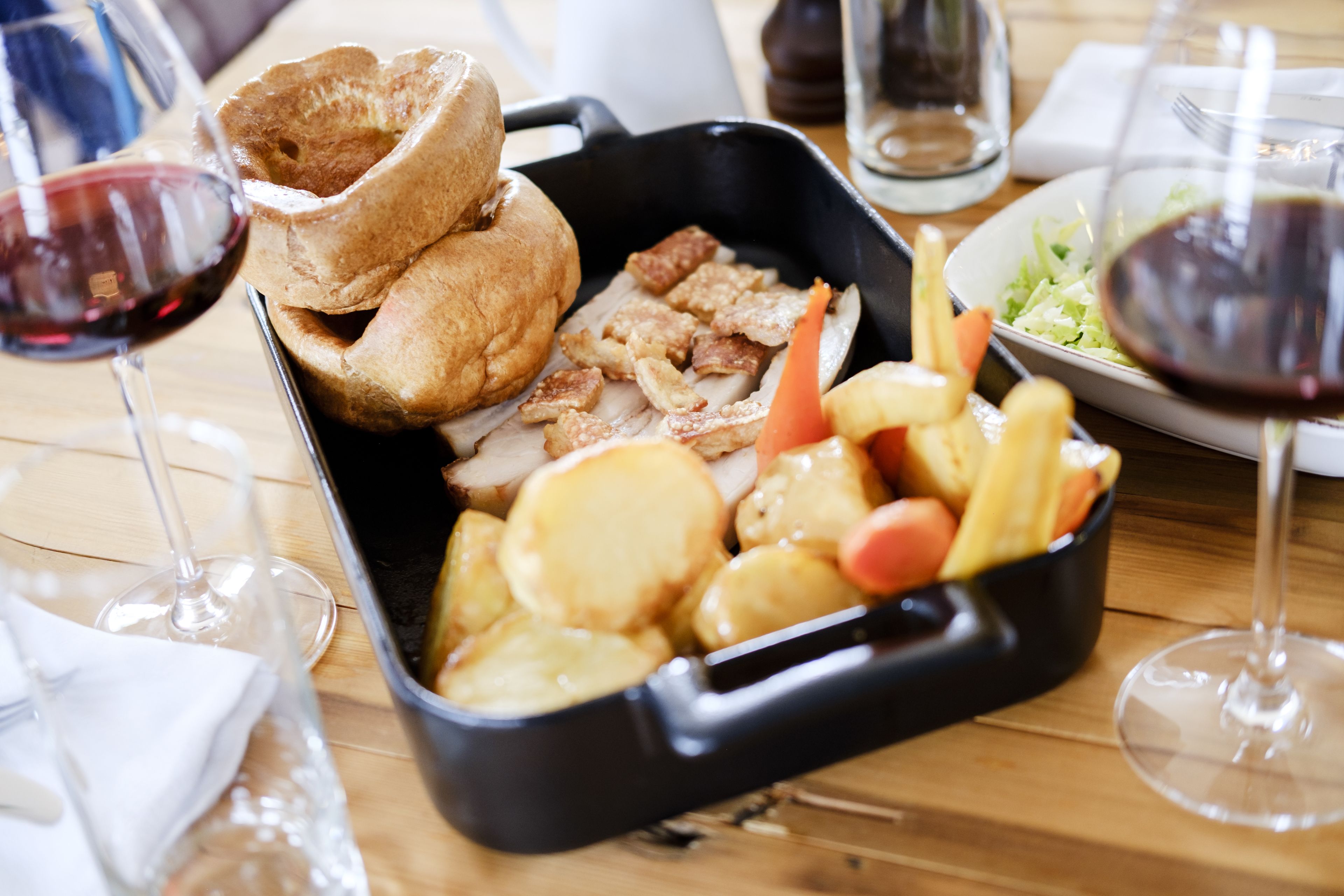 A meal with Yorkshire pudding, meat, and vegetables in a black pan, accompanied by wine glasses and a salad on a wooden table.