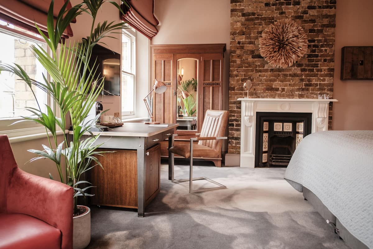 A bedroom with a brick wall, a fireplace, a bed, a desk, a chair, and a potted plant.