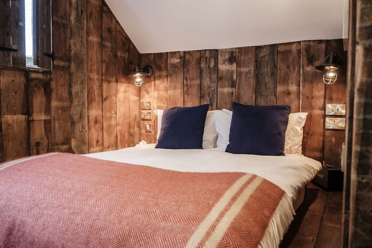 A bedroom with a wooden wall, a bed with a patterned blanket, two pillows, and two wall-mounted lights.
