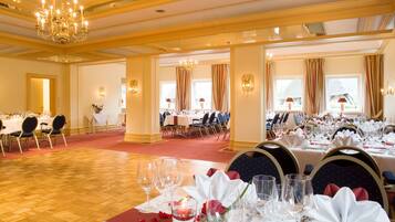 Un salón de banquetes con varias mesas redondas dispuestas para comer, candelabros y ventanas grandes con cortinas.