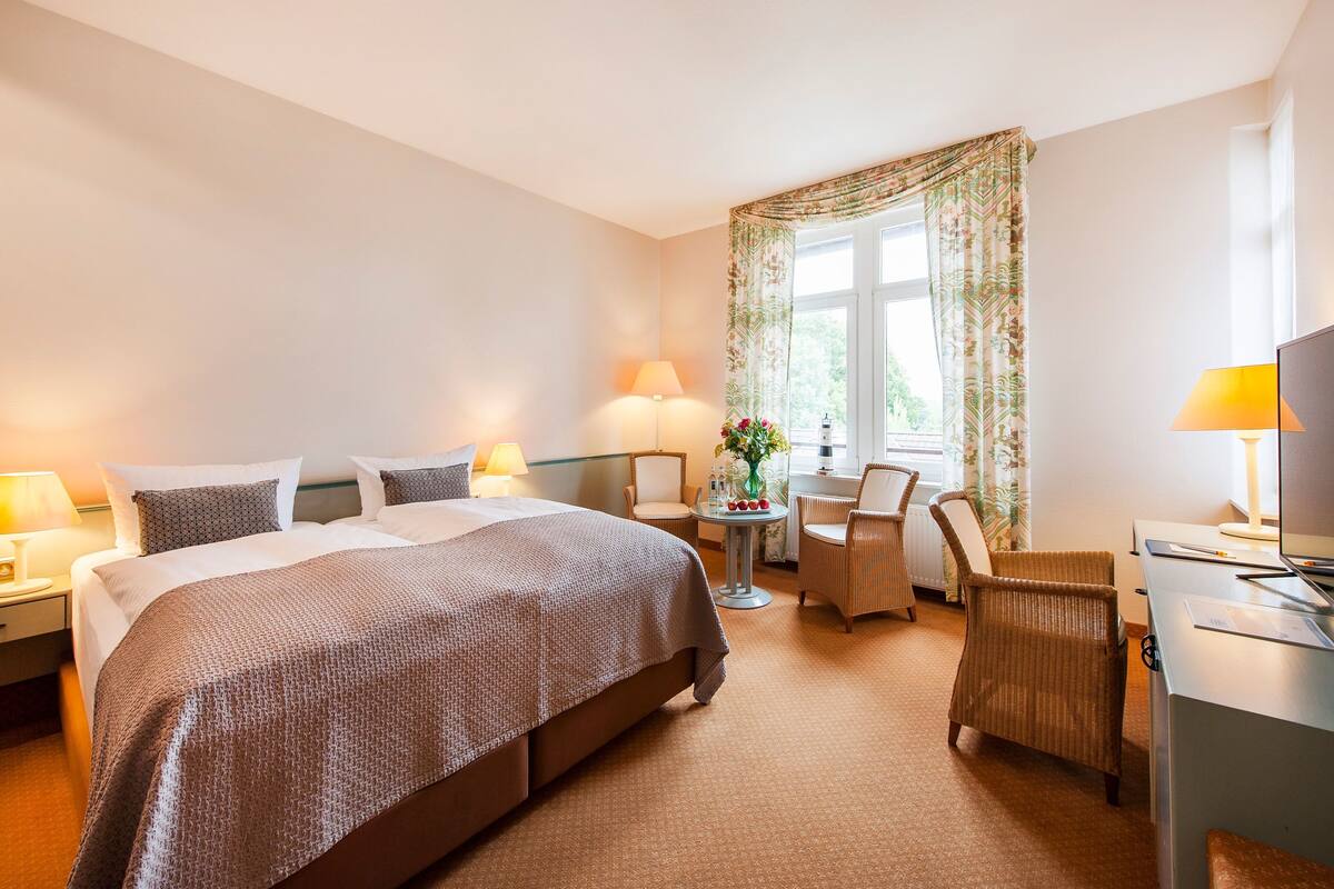 Una habitación de hotel con una cama grande, dos mesitas de noche con lámparas, un escritorio con una silla, una pequeña mesa redonda con un florero y una ventana con cortinas.
