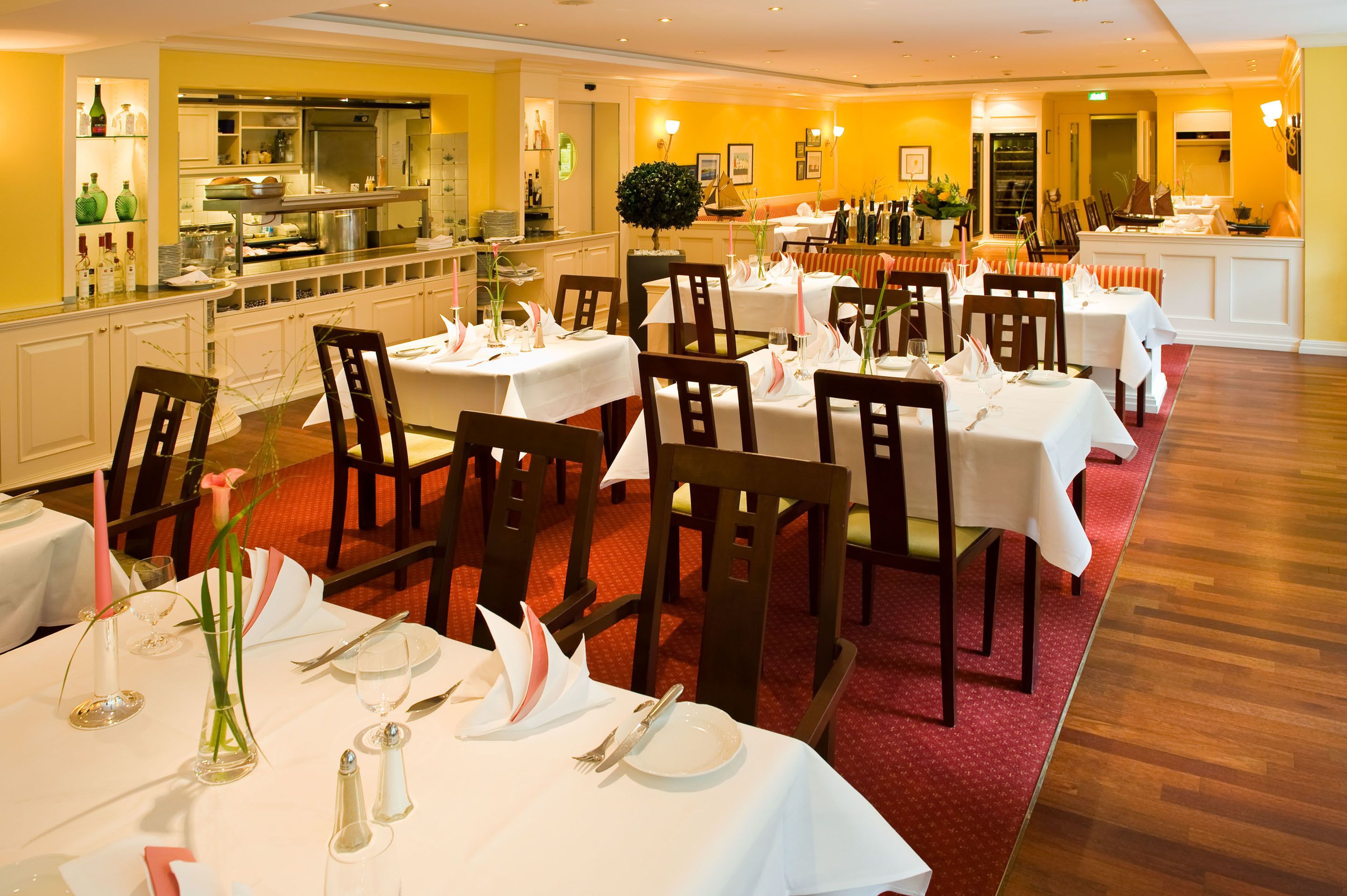 Un restaurante con varias mesas dispuestas para comer, una zona de cocina y una alfombra roja que lleva al área de asientos.