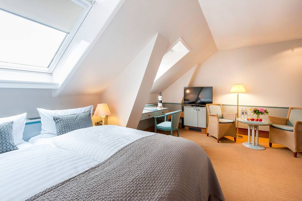 Una habitación de hotel moderna con techo inclinado, una cama con ropa de cama blanca, un escritorio con una silla, un televisor y una mesita de noche con lámpara y flores.