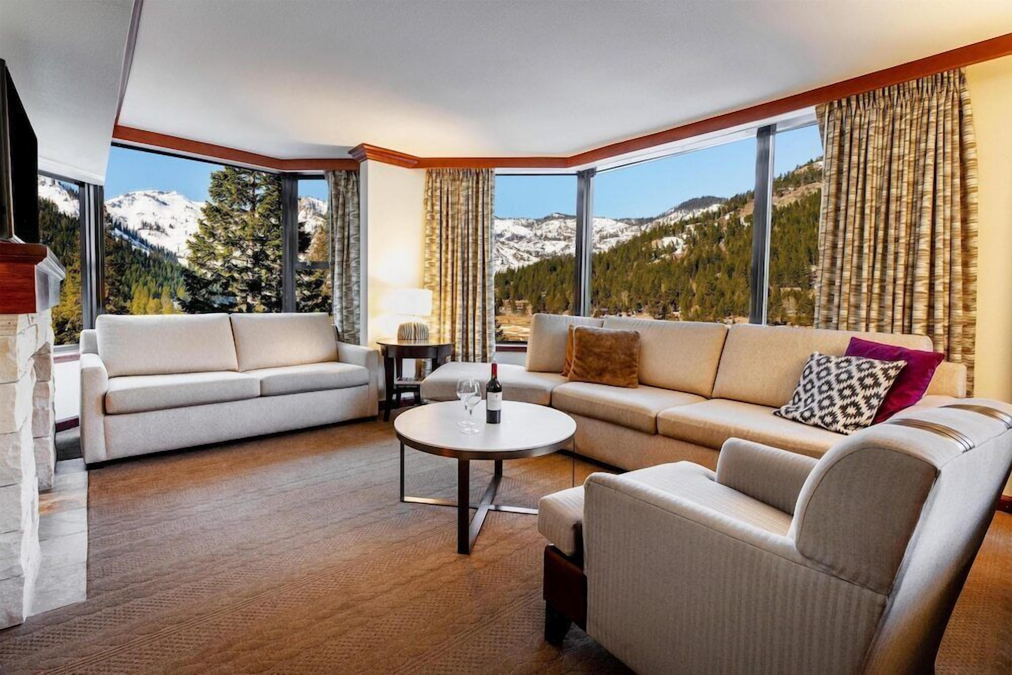 A modern living room with a sofa, armchair, and coffee table, large windows with mountain views, and a stone fireplace.