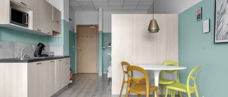 A modern kitchen with wooden cabinets, a microwave, and a coffee machine. There is a dining area with a white table and yellow chairs.