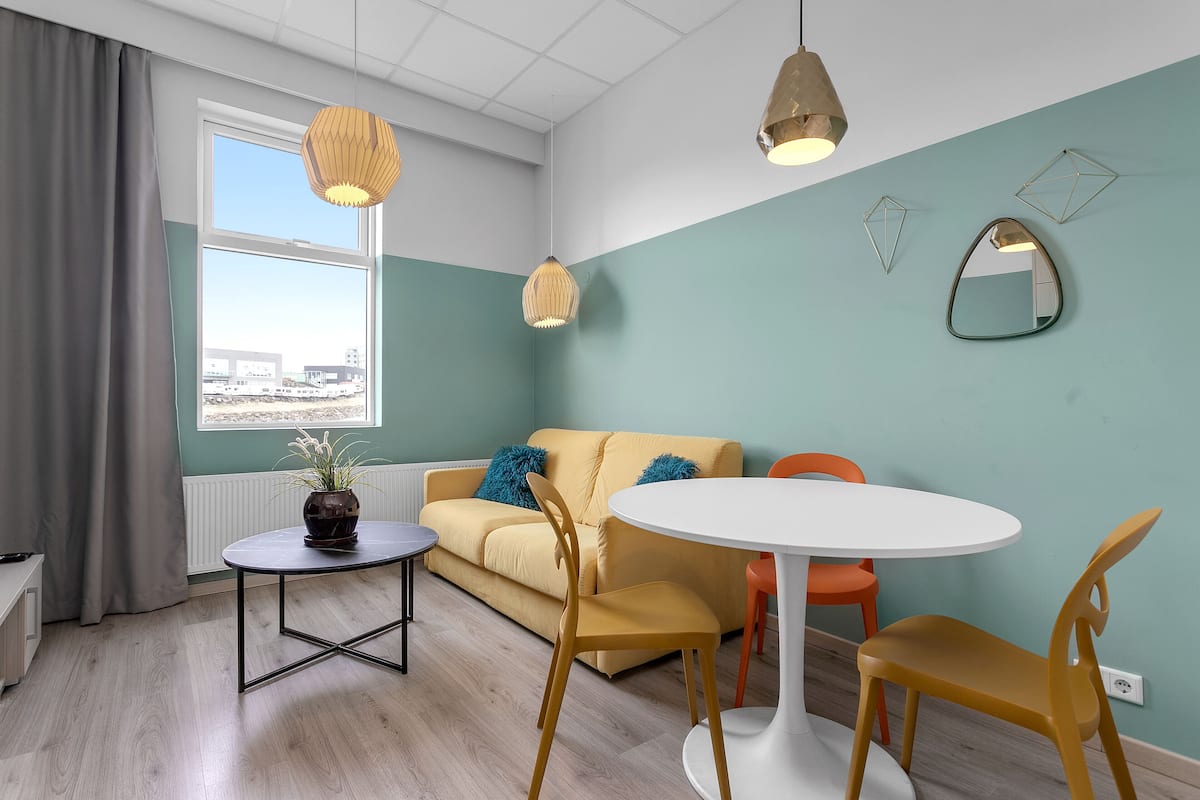 Un salon moderne avec un canapé, une table ronde et deux chaises. Une fenêtre offre une vue sur les bâtiments environnants.