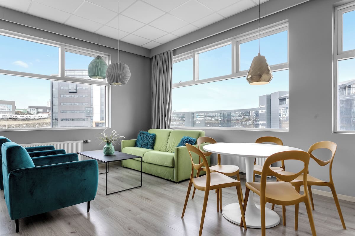 Un salon moderne avec un canapé vert, des fauteuils turquoise et des chaises en bois disposées autour d’une table blanche. De grandes fenêtres offrent une vue sur la ville.