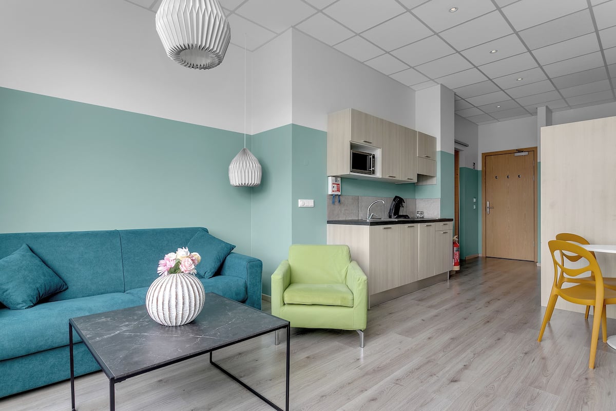 Un salon moderne avec un canapé turquoise, un fauteuil vert, une table basse en marbre et un coin cuisine doté de meubles en bois et d’un four à micro-ondes.