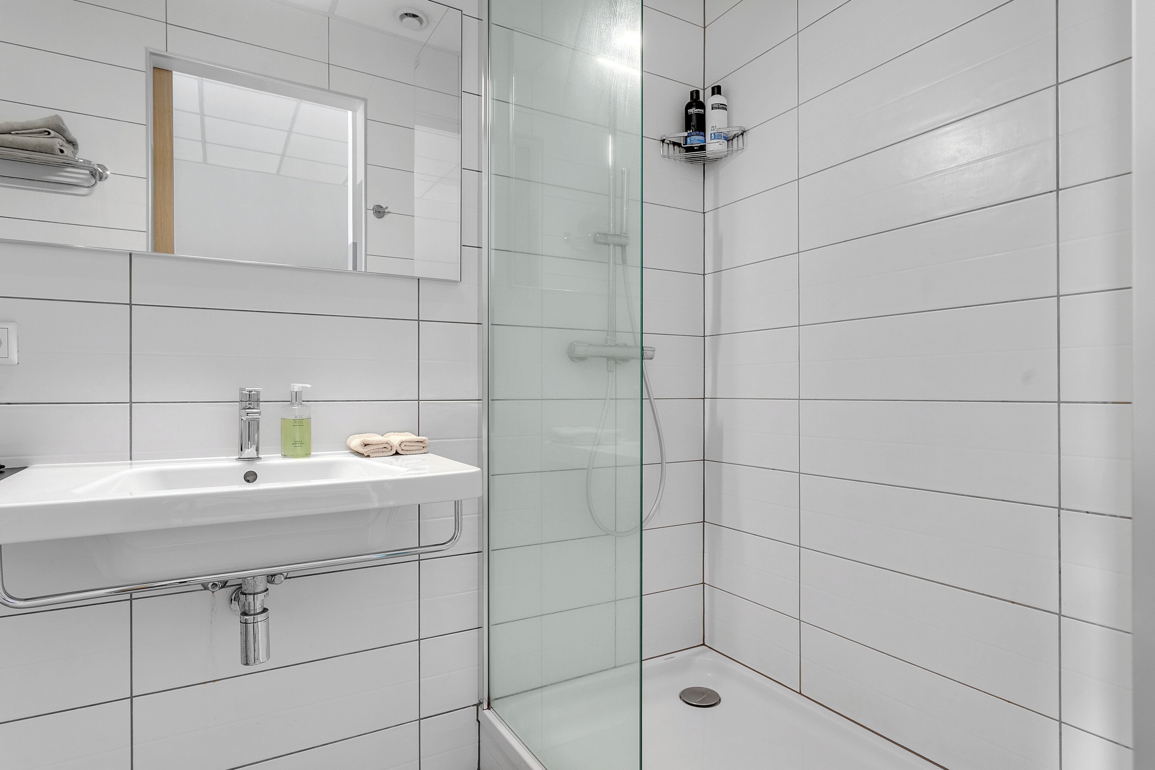 Un baño moderno con ducha de vidrio, un lavabo blanco con espejo y un estante con artículos de tocador.