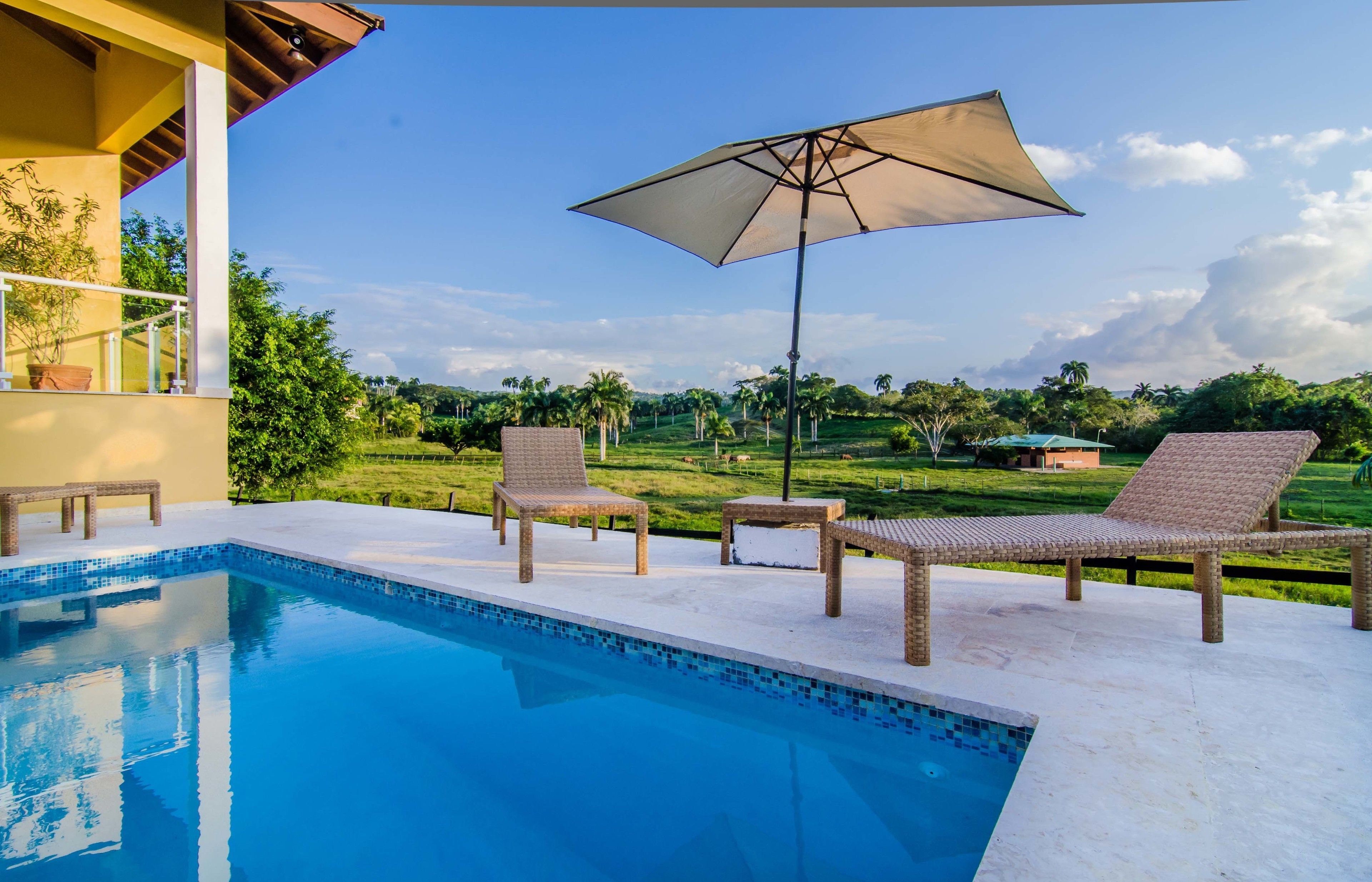 A poolside area with lounge chairs and an umbrella, surrounded by a lush landscape and a clear sky.