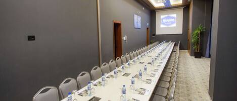 A conference room with a long table, chairs, water bottles, and a projector screen displaying "Hampton by Hilton".