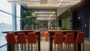 A modern restaurant with a long communal table and orange chairs, surrounded by large windows and a ceiling with linear light fixtures. - Hampton by Hilton Bolu (Bolu)