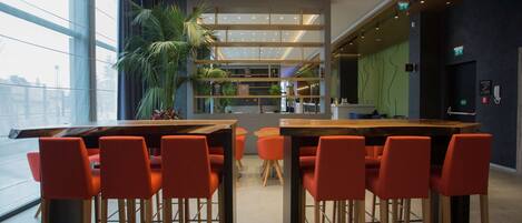 A modern restaurant with a long communal table and orange chairs, surrounded by large windows and a ceiling with linear light fixtures.