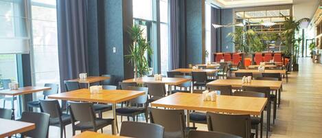 A spacious dining area with multiple tables and chairs, large windows, and indoor plants.