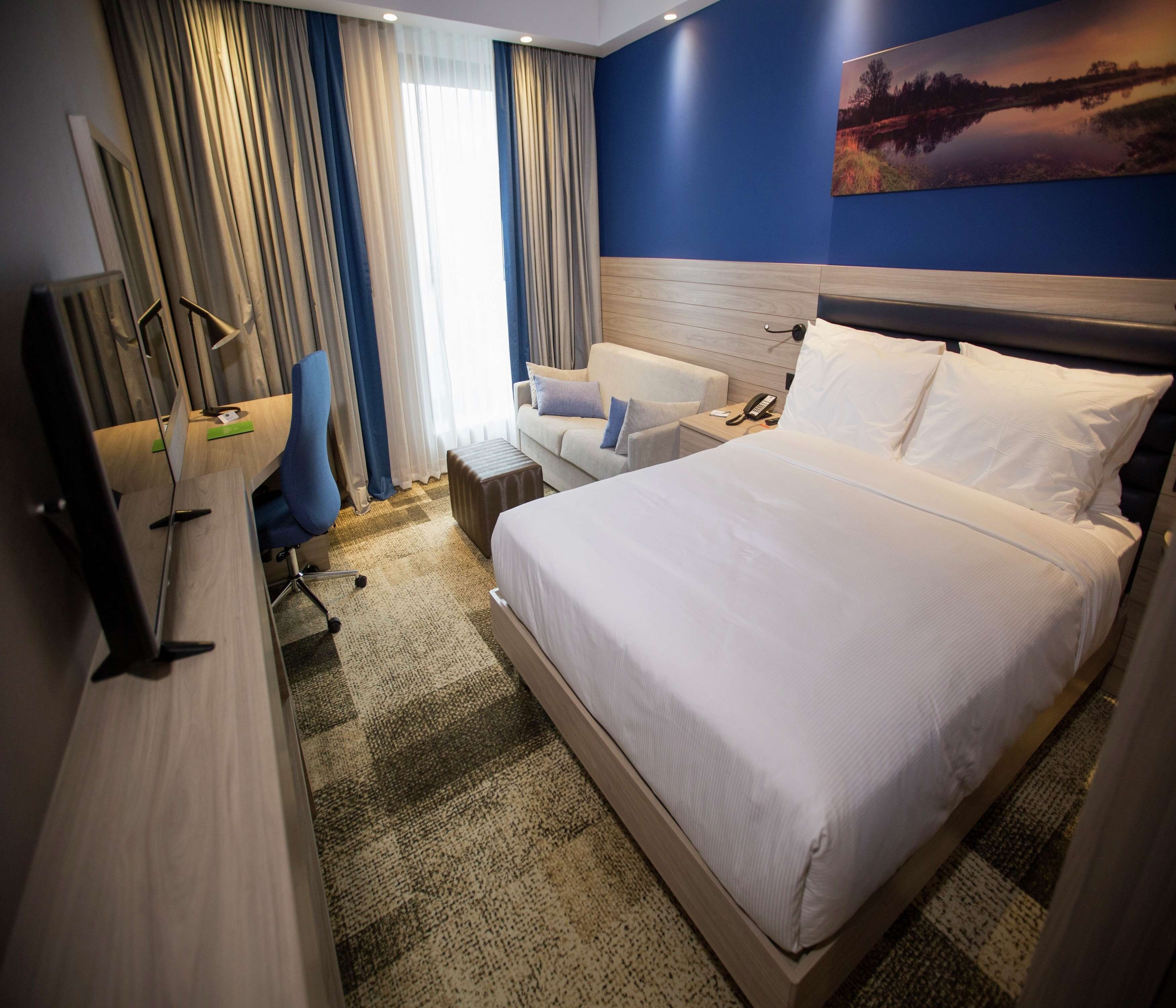 A hotel room with a large bed, a desk with a chair, a small table, and a painting on the wall.
