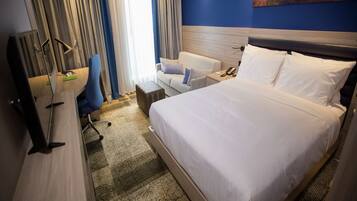 A hotel room with a large bed, a desk with a chair, a small table, and a painting on the wall.