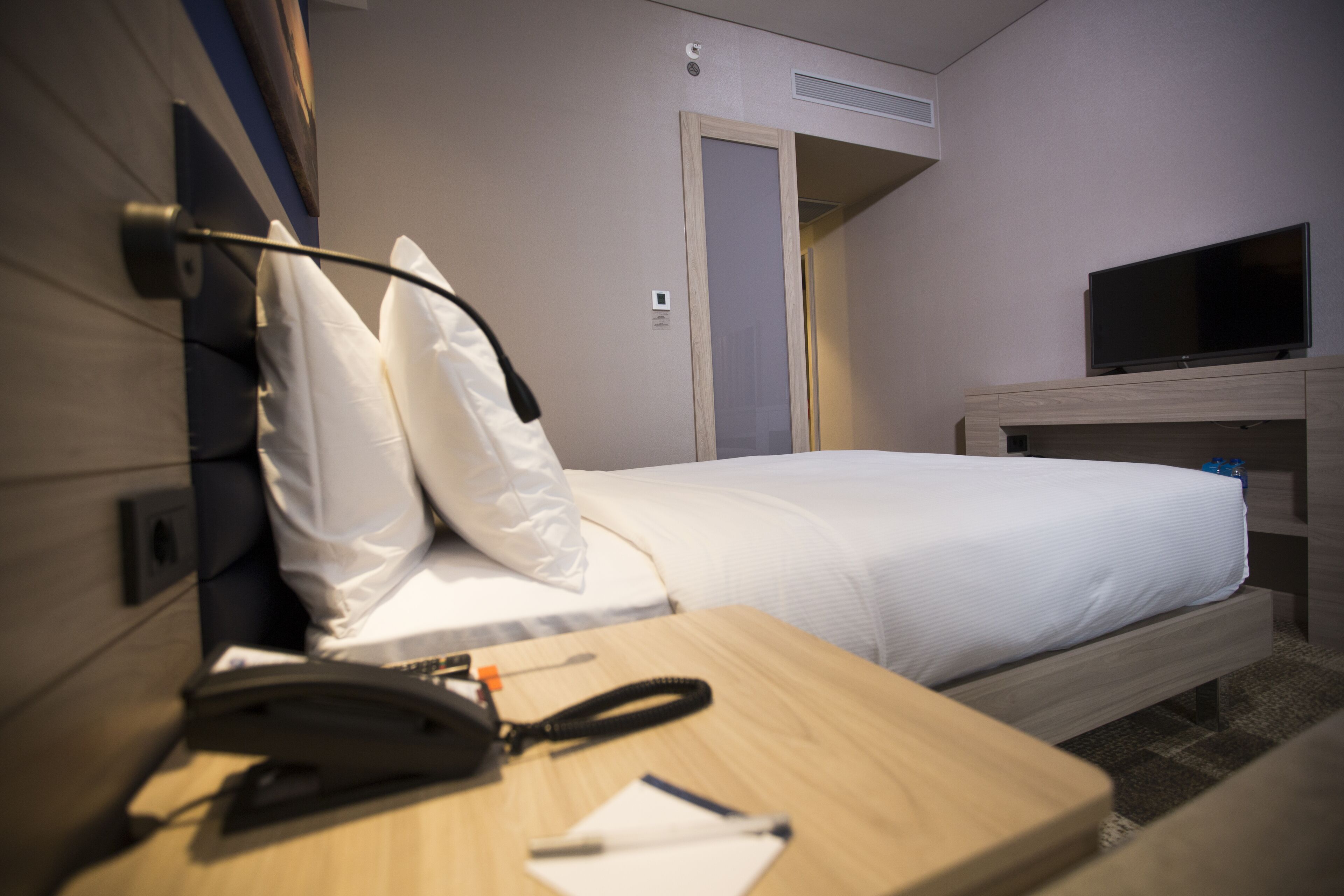 A hotel room with a bed, a desk with a telephone, a television on a stand, and a door leading to another room.