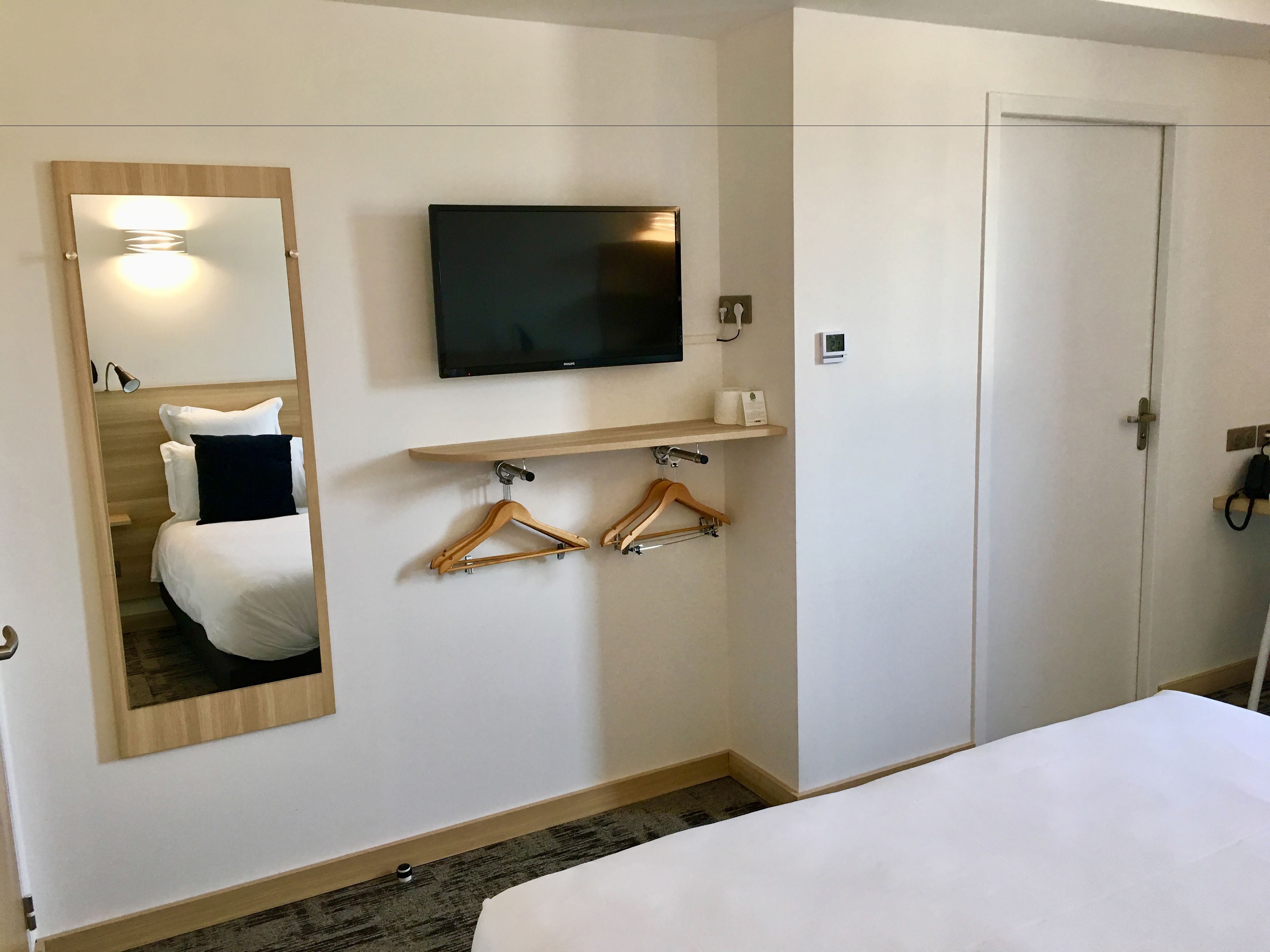 Habitación de hotel con cama, televisor montado, espejo, perchero y puerta.