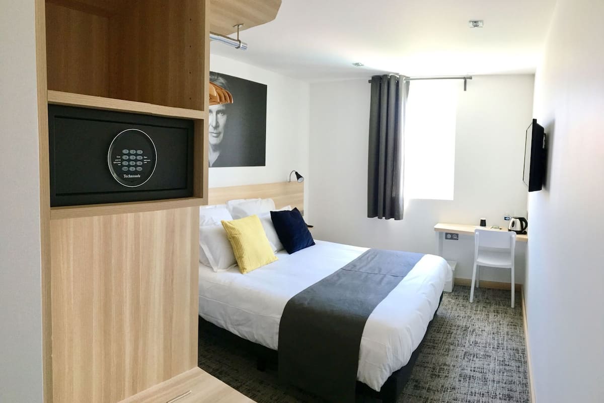 Una habitación de hotel con cama, escritorio, silla, televisión, ventana con cortinas y panel de control en la pared.
