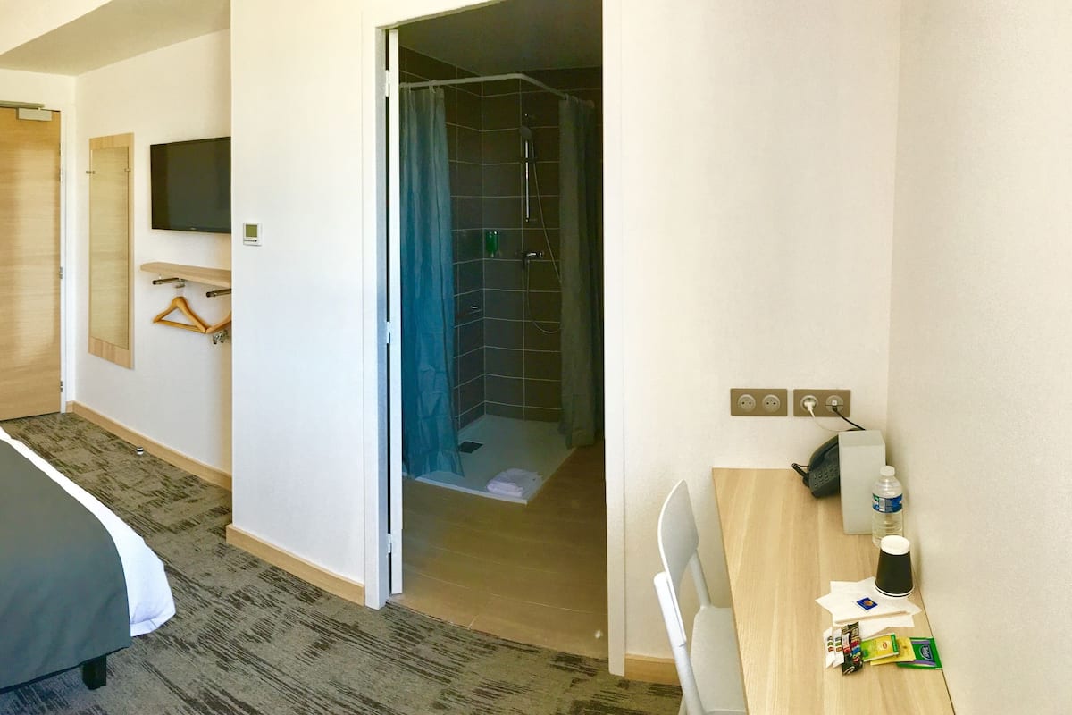 Habitación de hotel con cama, televisión, ducha y escritorio con artículos de aseo.