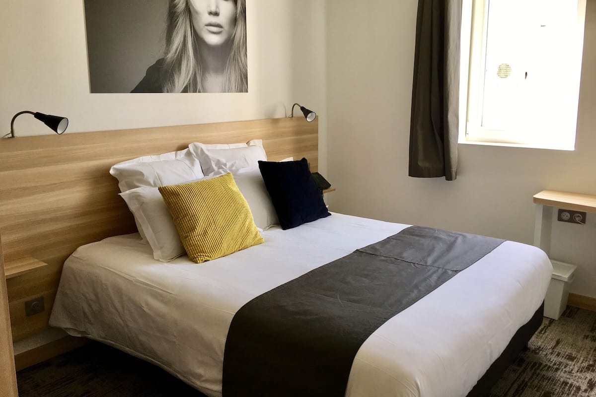 Una cama bien hecha con almohadas blancas y oscuras, un plaid gris y un cabecero de madera.