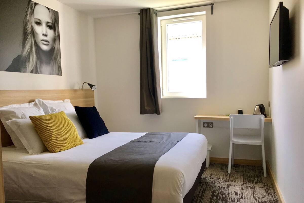 Habitación de hotel con cama, escritorio, silla, televisor, ventana con cortinas e imagen colgada en la pared.