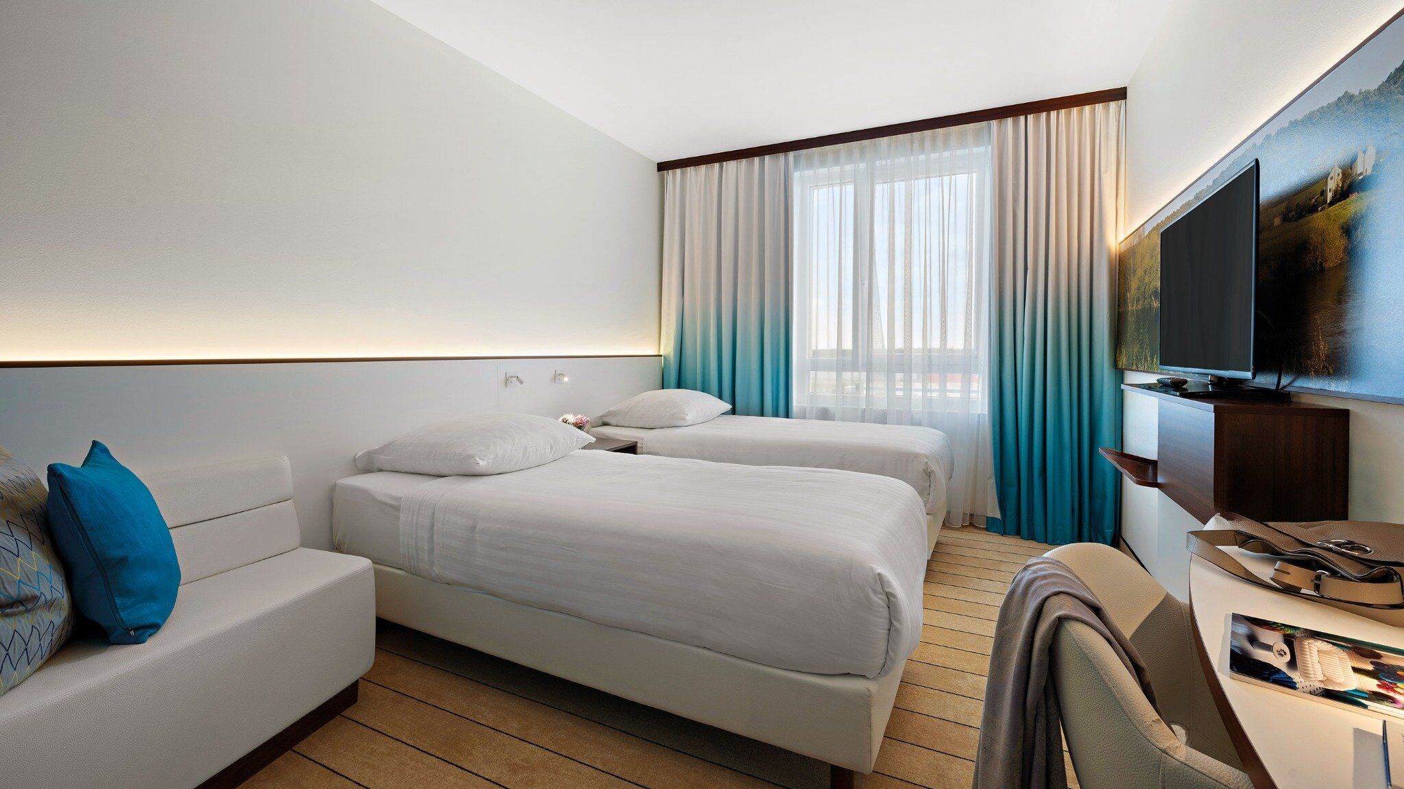 A hotel room with two beds, a sofa, a desk with a TV, and a large window with curtains.