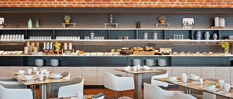 Um balcão de pequeno-almoço moderno com parede de tijolos, espaço para buffet e área de refeições com cadeiras e mesas brancas.