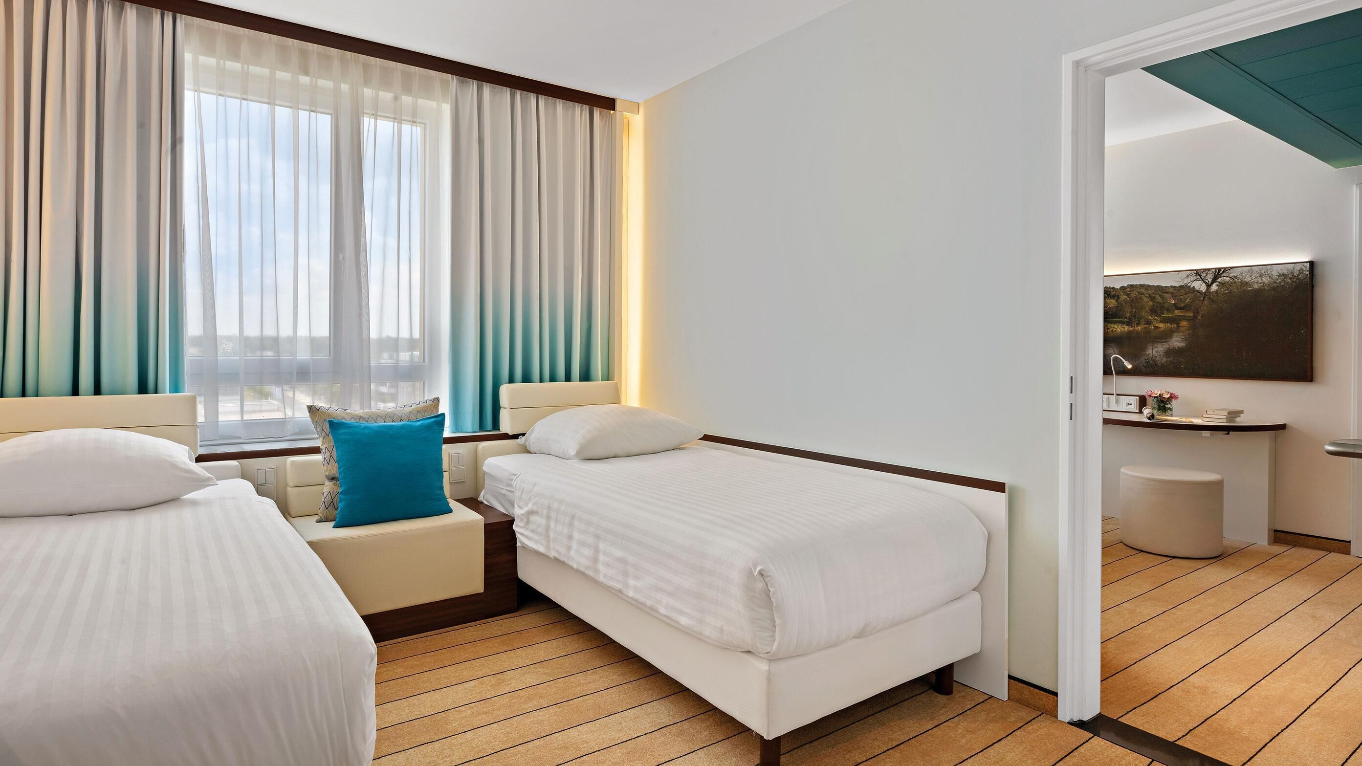 A hotel room with a bed, a bedside table, a chair, a window with curtains, a painting on the wall, and a small stool.