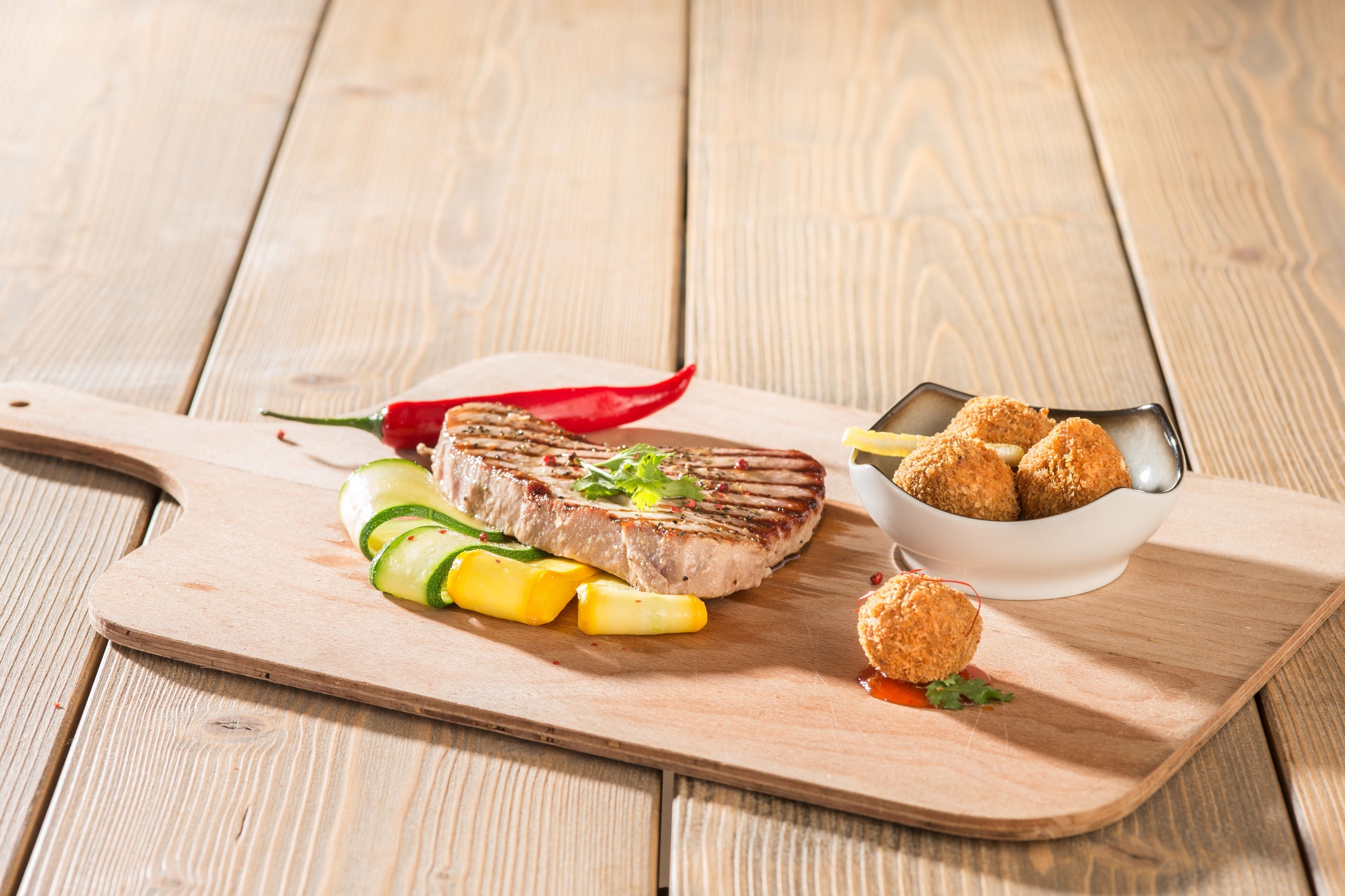 A wooden cutting board with a grilled steak, sliced zucchini, pineapple, and a red chili pepper.