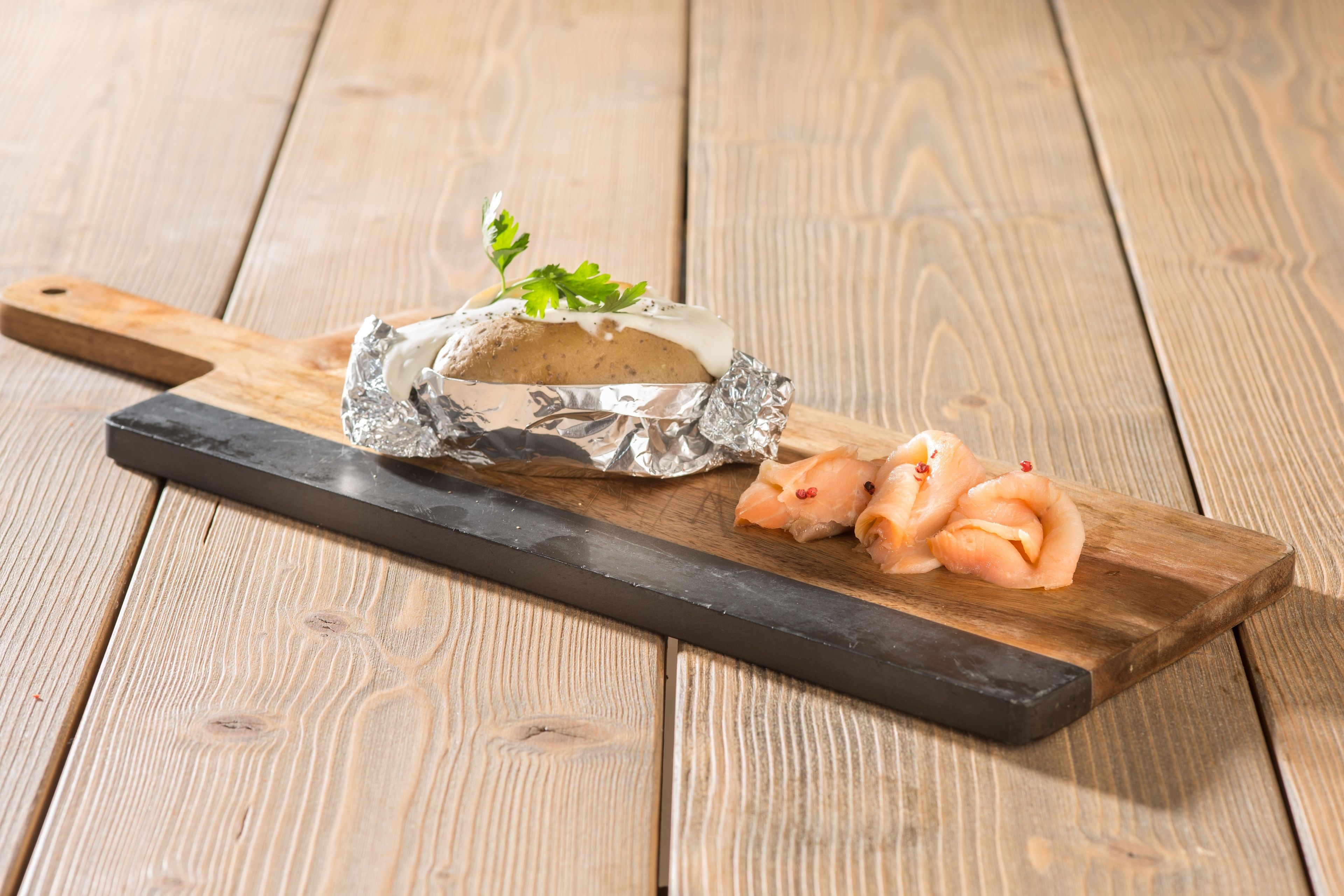 A wooden cutting board with a piece of food wrapped in foil and sliced food on a wooden surface.
