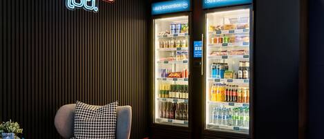 A modern lounge area with a neon sign, two refrigerated display cases, a grey sofa, and a small table with a plant.