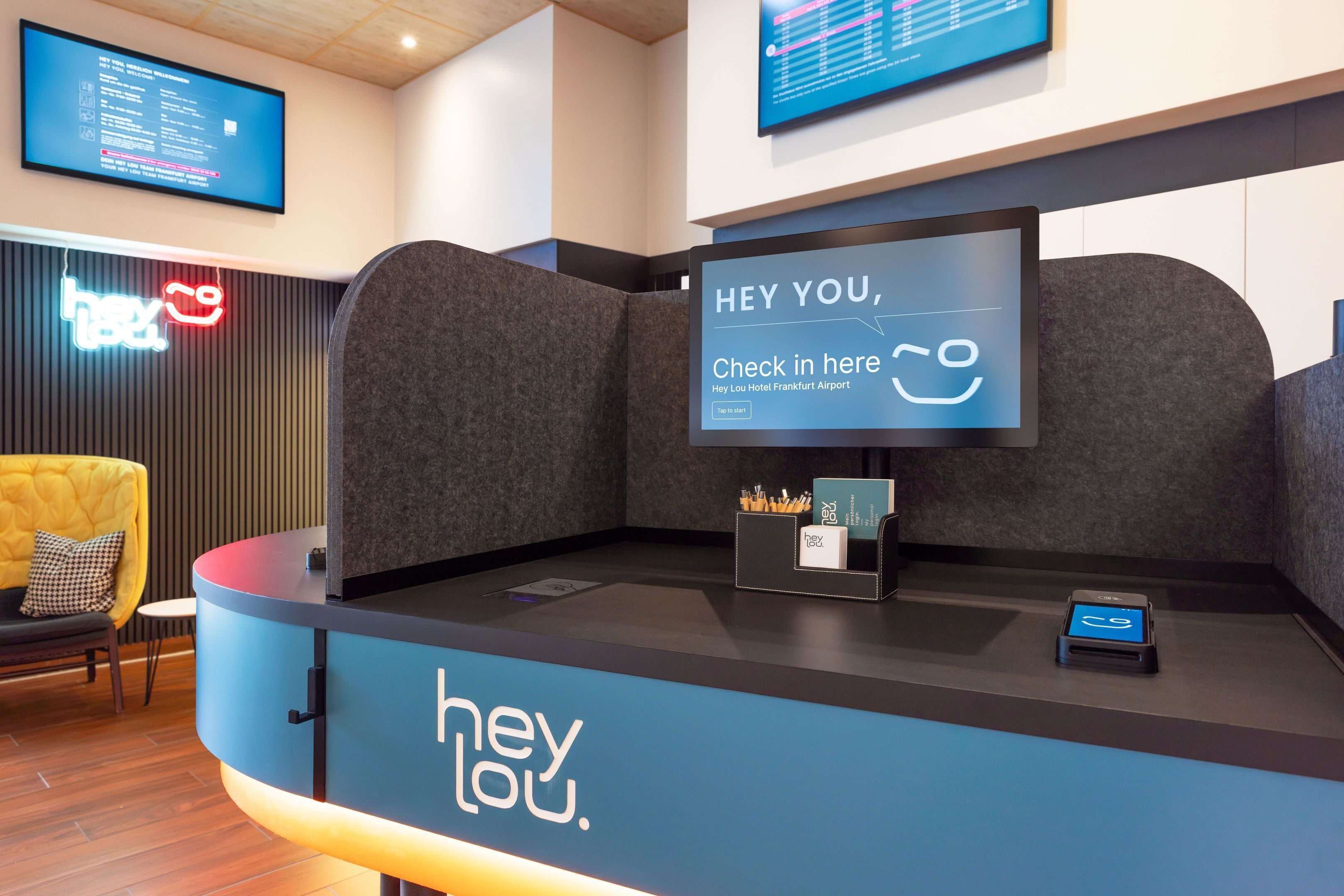 A reception desk with a digital screen displaying a check-in message, a neon sign, and a seating area with a yellow chair.