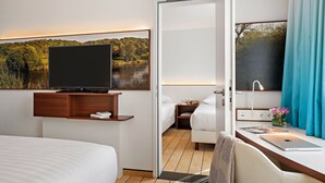 Habitación de hotel con cama, escritorio con computadora portátil, televisor en estante de madera y ventana con vista a un lago y árboles.