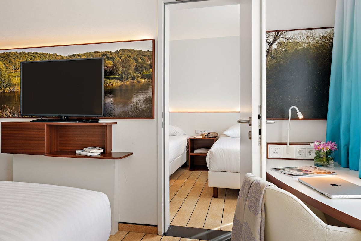 Quarto de hotel com uma cama, uma secretária com um laptop, uma TV numa estante de madeira e uma janela com vista para um lago e árvores.