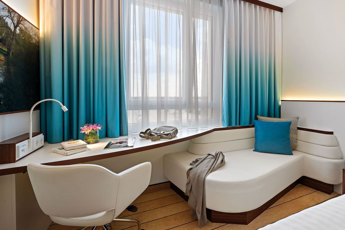 Quarto de hotel com escrivaninha, cadeira, cama e janela com cortinas.
