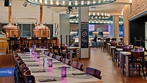 A restaurant with long communal tables, wooden chairs, and a bar area with a bartender. - Hey Lou Hotel Frankfurt Airport (Kelsterbach)