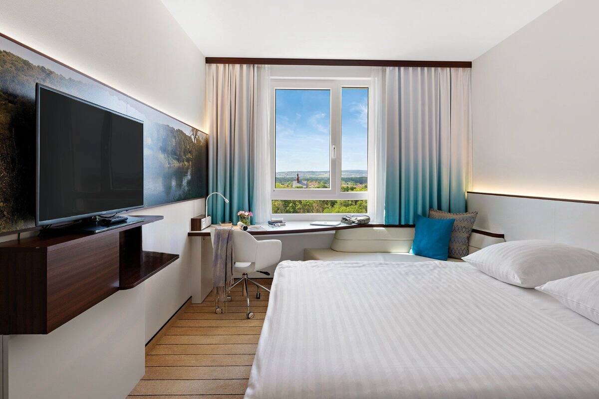 Quarto de hotel com cama, escrivaninha com cadeira, televisão e janela com cortinas.